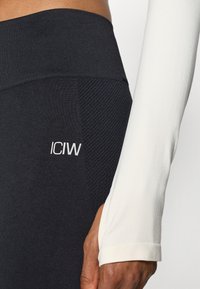 Μαύρες κολάν με υφασμάτινες πάνελ, ψηλή μέση και λευκό λογότυπο. Λευκό μπλουζάκι με μακριά μανίκια που διαθέτει εφαρμοστό μανίκι.