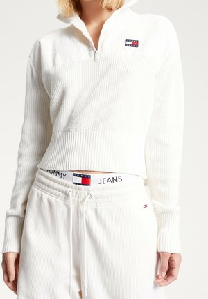 Kobieta ubrana w biały sweter z półzamkiem i prążkowanym wzorem oraz dopasowane spodnie, z widocznym logo Tommy Jeans na pasku spodni i na piersi swetra.