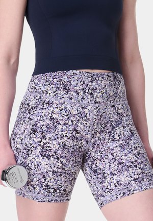 Vrouw draagt hoge taille sportshorts met bloemenpatroon en een marineblauw crop top, houdt een chrome dumbbell van 3 kg in haar rechterhand vast.