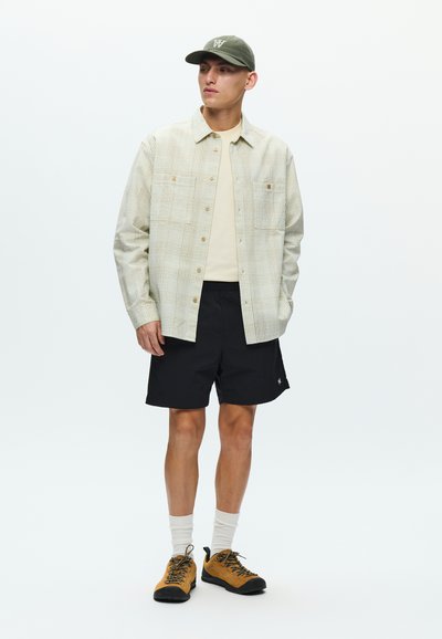 Chemise à carreaux claire boutonnée, t-shirt beige, short noir et chaussures de randonnée jaunes. Casquette verte et chaussettes blanches complètent la tenue.
