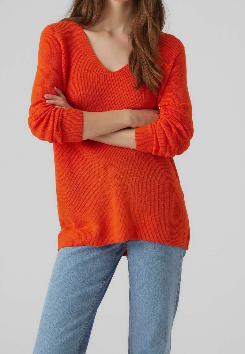 Pull en maille orange vif avec un décolleté en V profond, des manches longues et une texture côtelée, associé à un jean bleu clair.