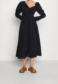 Svart långärmad klänning med djup V-ringning, veckad byst och ett band i midjan. Matchad med bruna läderklackar. Slät textur; ankel-lång design.