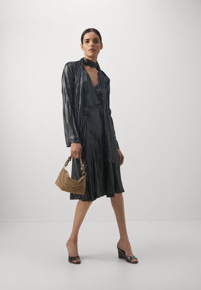 Robe noire plissée avec un décolleté plongeant, assortie à un sac tissé beige et des mules à talons transparents. Texture de tissu lisse et brillant.