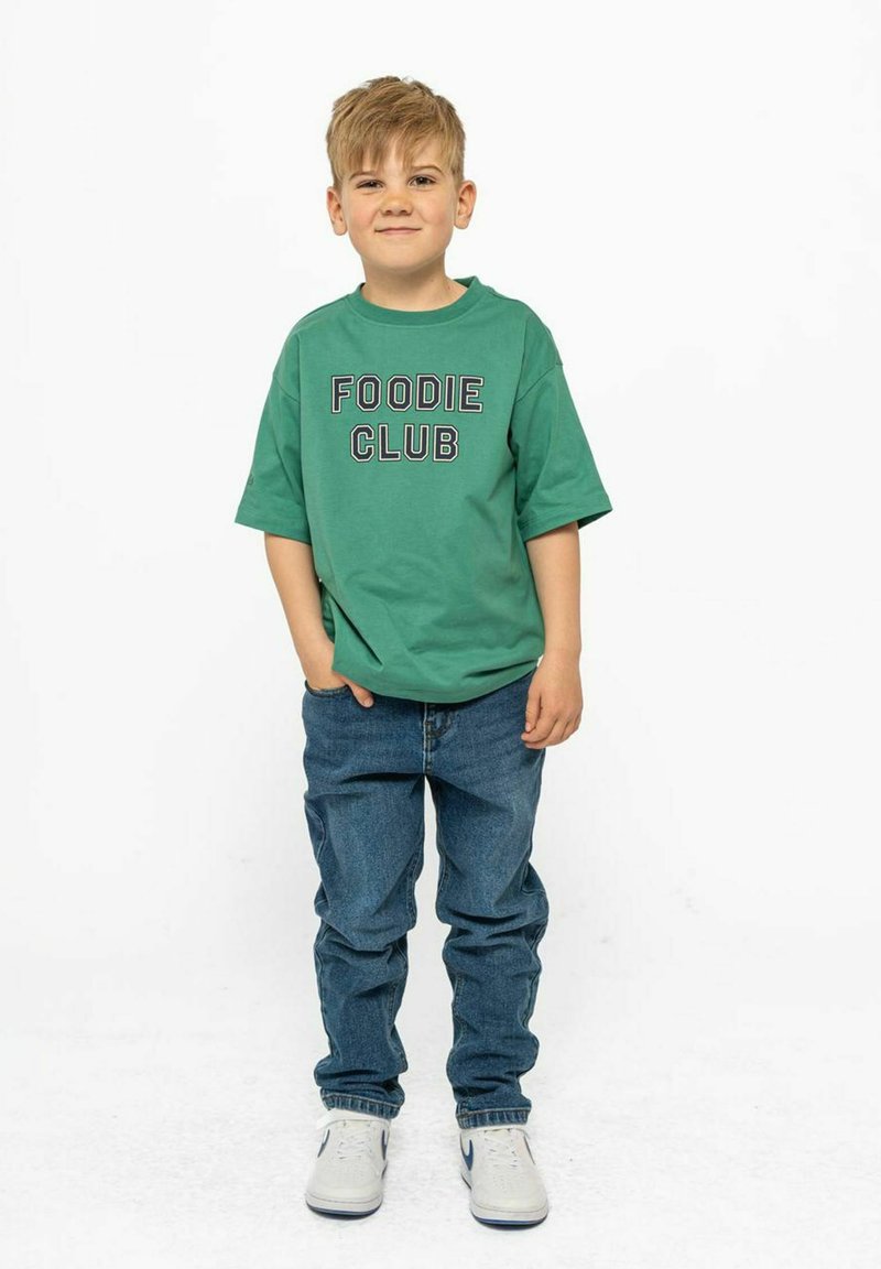 Zelena pamučna majica s natpisom "FOODIE CLUB" ispisanim crnom bojom, u kombinaciji s plavim trapericama i bijelim tenisicama s plavim akcentima.