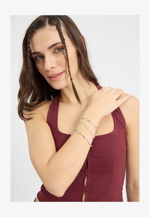 Sudraba rokassprādzes ar dažādiem dizainiem, tostarp sirdīm un ķēdēm, uz rokas. Augšdaļa ir burgundijas krāsā ar kvadrātveida kakla izgriezumu un pogu detaļām.