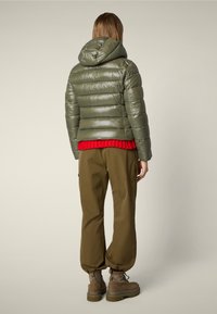 Giacca a vento verde oliva con cappuccio, finitura lucida e cuciture orizzontali, indossata sopra un maglione di lana rosso e abbinata a pantaloni marroni.