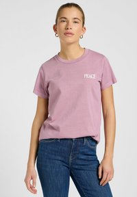 Camiseta de algodón rosa de manga corta y cuello redondo; presenta la palabra "PAZ" impresa en blanco en la parte superior izquierda. Llevada con jeans azules.