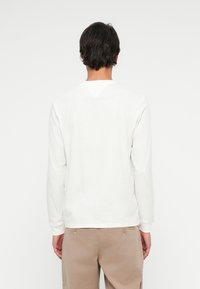 Mand iført en ensfarvet hvid langærmet skjorte og beige bukser, der står med ryggen til kameraet mod en hvid baggrund.