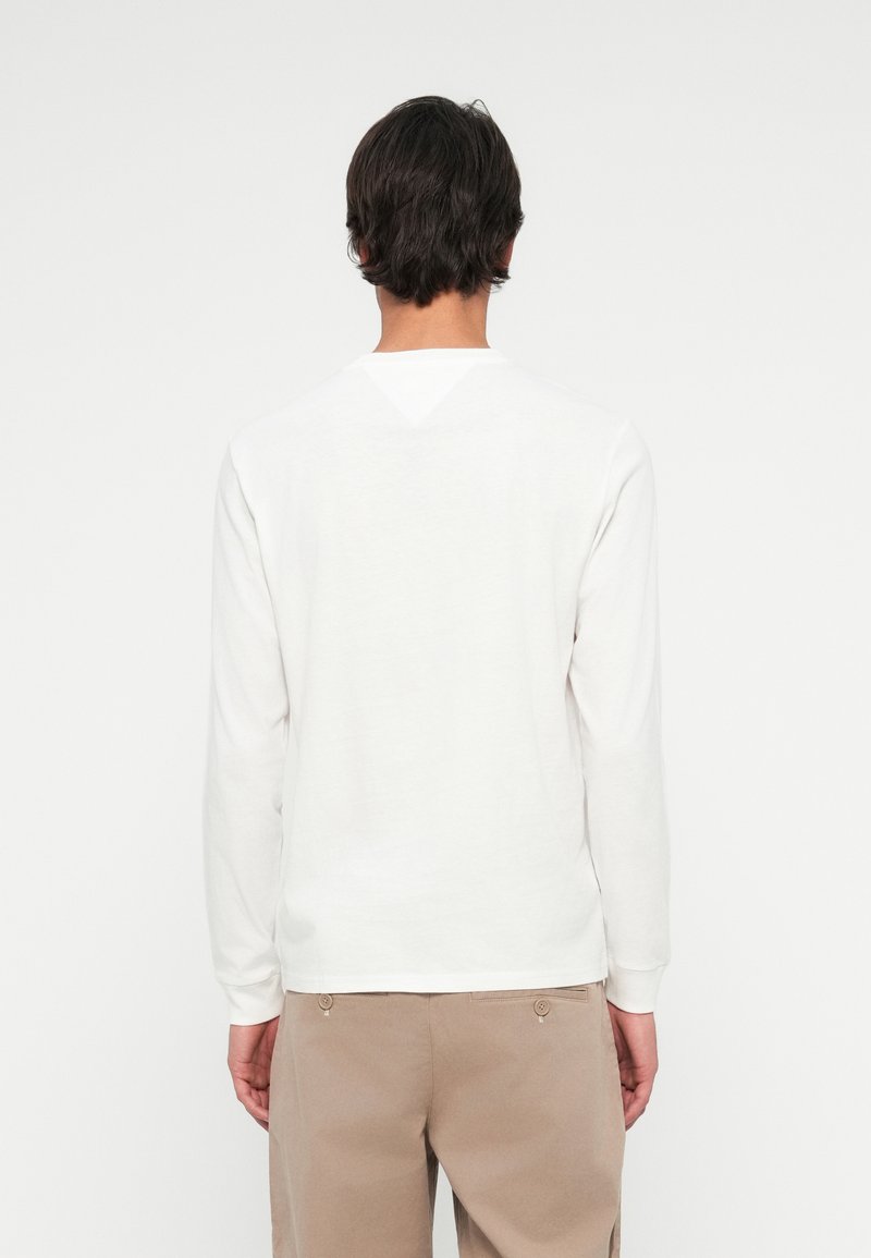 Hombre vestido con una camiseta blanca de manga larga y pantalones beige, de espaldas a la cámara contra un fondo blanco.