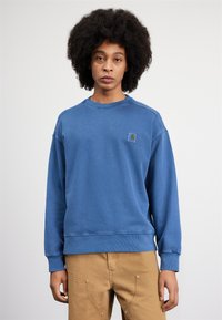Sudadera de forro polar azul con cuello redondo, puños acanalados y un logo de parche en el lado izquierdo del pecho. Material de algodón con una textura suave.