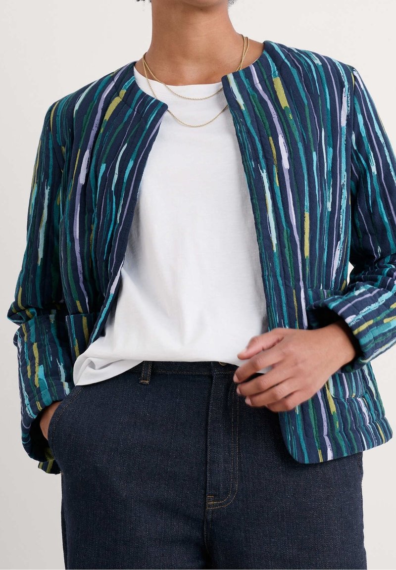 Kurtka pikowana w pionowe niebieskie i zielone paski, z okrągłym dekoltem, długimi rękawami oraz otwartym przodem, noszona na białej koszuli.