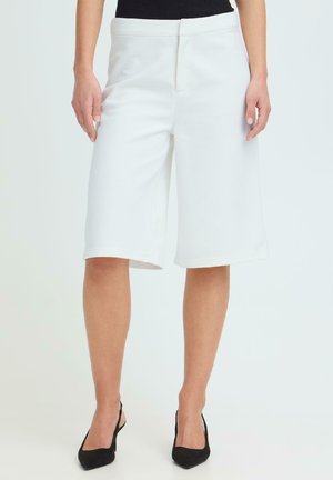 Vrouw draagt witte op maat gemaakte shorts tot op de knie en zwarte slingback pumps, staand tegen een effen lichte achtergrond.