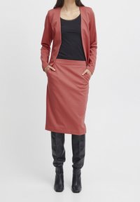 Rosa Blazer mit schlanker Passform, kombiniert mit einem schwarzen ärmellosen Oberteil und einem passenden mid-length Rock. Schwarze kniehohe Absatzstiefel.