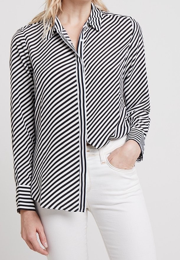 Mannequin portant une chemise boutonnée à rayures noires et blanches et un pantalon blanc, une main dans une poche.