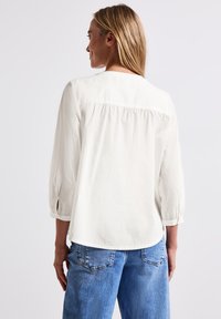 Weiße, leichte Bluse mit gerafftem Rückenteil und 3/4-Ärmeln. Mit Schlaufenakzenten an den Ärmelöffnungen. Kombiniert mit hellblauen Jeans.