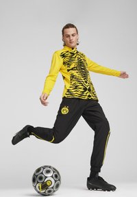 Maglietta sportiva a maniche lunghe gialla e nera con motivo a righe, abbinata a pantaloni neri con accenti gialli e logo, sopra un pallone da calcio a motivi.