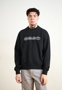 Sudadera negra con cuello redondo, mangas largas y puños de canalé. Presenta un logo ovalado blanco con las letras "CA RH AR TT" en el pecho.