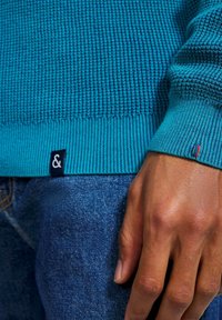 Ingrandimento di una mano appoggiata su jeans blu, che indossa un maglione texture teal con polsini a costine e un piccolo etichetta nera in tessuto.