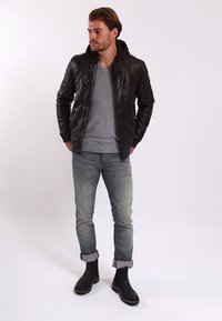 Giubbotto di pelle marrone con cappuccio, maglione grigio, jeans in denim azzurro chiaro e stivaletti neri alla caviglia. Modello in piedi contro uno sfondo bianco.