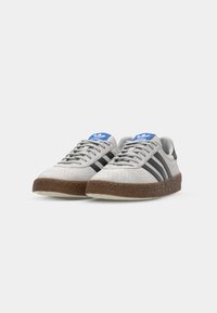 Grå suede sneakers med brune teksturerede gummisåler, der har tre sorte striber og et blåt adidas-logo på tungen.