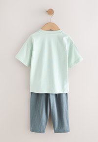 Chemise vert clair à manches courtes et short gris jusqu’au genou suspendus sur un cintre en bois contre un mur beige.
