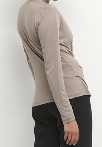 Langärmliges Oberteil aus leicht beigem Stoff mit glatter Textur, das über ein figurbetontes Design und dezente Drapierungen an den Seiten verfügt. Kombiniert mit schwarzen Hosen.