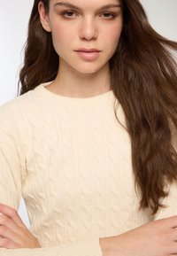 Młoda kobieta z długimi brązowymi włosami, ubrana w sweter z warkoczowym splotem w kolorze kremowym, z rękami skrzyżowanymi na piersi, patrząca prosto przed siebie z neutralnym wyrazem twarzy.