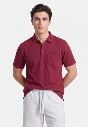 Junger Mann mit dunklen Haaren, der ein burgunderfarbenes Polohemd mit kurzen Ärmeln und hellgraue Shorts mit Kordelzug trägt und mit den Händen hinter dem Rücken steht.