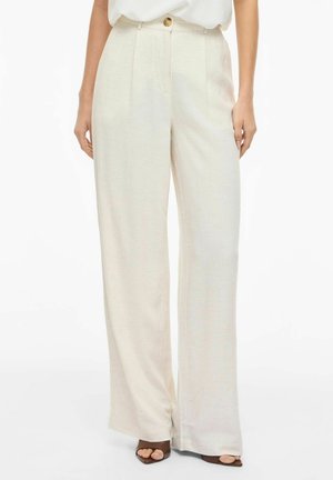 Kobieta w beżowych lnianych spodniach o wysokim stanie i szerokich nogawkach z guzikami oraz ciemnych butach na obcasie z odkrytymi palcami, stojąca na tle białego tła.