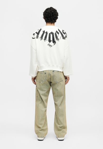 Άτομο με σγουρά μαλλιά στέκεται με την πλάτη, φοράει άσπρο φούτερ με μαύρο κείμενο "Angels" στην πλάτη και φαρδιά ανοιχτό μπεζ τζιν.