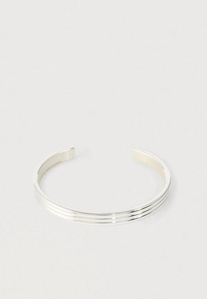 Strieborný náramok na zápästie s tromi paralelnými drážkami, otvorený dizajn, položený na bielej ploche.