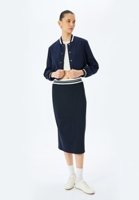 Veste de bombardier marine avec des rayures blanches, longueur cropped et boutons-pression, assortie à une longue jupe marine avec une taille rayée et des baskets blanches.