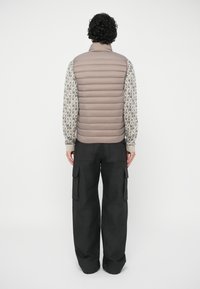 Muškarac s kovrčavom kosom nosi bež kviltani prsluk preko dugih majica s uzorkom i labavih crnih cargo hlača, stoji naslonjen uz jednostavnu bijelu pozadinu.