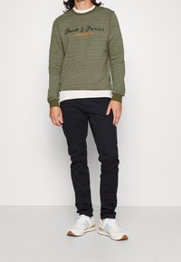 Felpa verde oliva con una finitura testurizzata, caratterizzata da un logo e dalla scritta 'Established 1990' in arancione, abbinata a pantaloni neri slim-fit e sneakers.