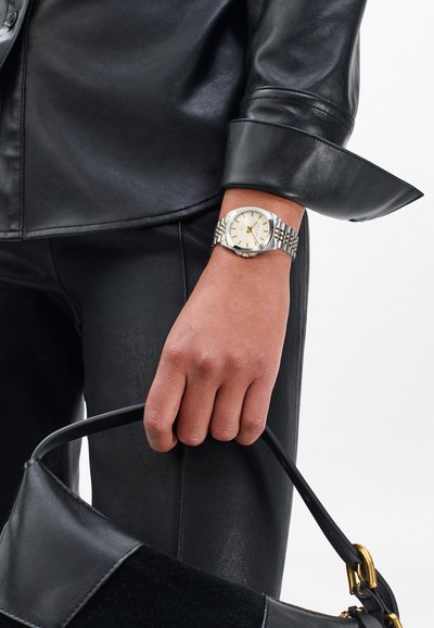 Sølv- og guldkolærur med hvid urskive og metallisk rem, kombineret med en sort læderskjorte og en sort taske med gulddetaljer.