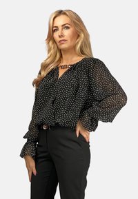 Schwarze Bluse mit Punkten, losen, durchsichtigen Ärmeln und einem Schlüssellochausschnitt. Kombiniert mit schwarzen Hosen und einem dekorativen Gürtel.