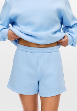 Persona che solleva una felpa azzurro chiaro mostrando l'addome, indossando pantaloncini a vita elastica coordinati dello stesso colore su uno sfondo semplice.