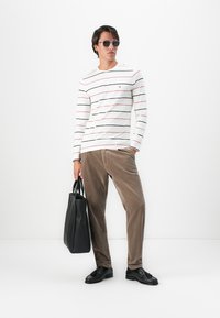 Uomo che indossa occhiali da sole, camicia a righe bianca a maniche lunghe, pantaloni di velluto a coste marroni, mocassini neri, mentre tiene una borsa a tracolla nera, in piedi contro un muro bianco.