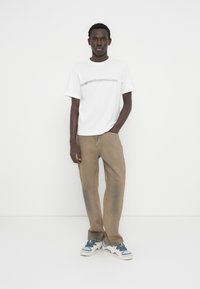 Λευκό κοντομάνικο μπλουζάκι με οριζόντια μαύρη ρίγα, συνδυασμένο με ανοιχτόκαφέ τζιν και γκρι αθλητικά παπούτσια, στέκεται μπροστά σε λευκό φόντο.