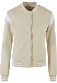 Veste universitaire beige avec des rayures blanches sur le col, les poignets et l'ourlet côtelés, des boutons-pression à l'avant et des accents blancs aux épaules.