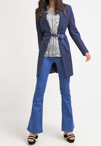 Marineblauer Gürteljacke über einem gemusterten Oberteil, kombiniert mit weit geschnittenen blauen Jeans und schwarzen High Heels. Glatter Stoff, taillierte Passform und strukturierte Gestaltung.