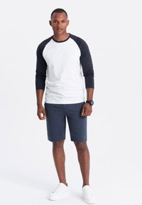 Maglietta a maniche raglan bianca e blu navy abbinata a pantaloni corti blu scuro con texture, completata da sneakers bianche, il tutto presentato su uno sfondo neutro.