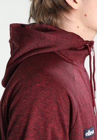 Maroon hooded sweatshirt with textured fabric, featuring a zippered neckline and logo patch on the left side. Drawstrings for hood adjustment.