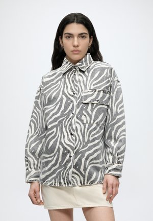 Γυναίκα με μακριά σκούρα μαλλιά που φοράει ένα γκρι και λευκό πουκάμισο με μοτίβο ζέβρας και μία ανοιχτό μπεζ φούστα, στέκεται μπροστά σε ένα απλό φόντο.