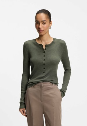 Kobieta w zielonym prążkowanym longsleeve Henley i beżowych spodniach, stojąca z jedną ręką w kieszeni na białym tle.