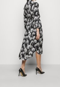 Černé šaty s bílým potiskem palmových listů, dlouhé rukávy, úzký pas a vysoký-nízký lem, doplněné černými stiletto podpatky.