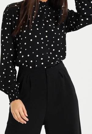 Kobieta w czarnej bluzce z długim rękawem w białe kropki i czarne spodnie z wysokim stanem, ręka oparta blisko biodra.