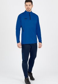 Top atletico a maniche lunghe blu con mezza zip e logo nero, abbinato a pantaloni della tuta blu navy con dettagli blu sui bottoni laterali.