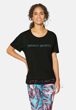 Lächelnde Frau mit lockigem Haar trägt ein schwarzes "Privacy Sports" T-Shirt und lebhaft blau-pink gemusterte Leggings und posiert mit der Hand in der Hüfte.