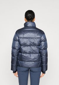 Navy blue puffer jacket with a high collar, quilted design, and shiny texture. Features a fitted silhouette and elastic cuffs.